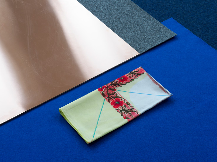 Materialmix auf dem Boden liegens: Kupferplatte, darunter grauer und blauer Filz, sowie eine bezeichnete Stick-Tischdecke. Ausstellungsdetail