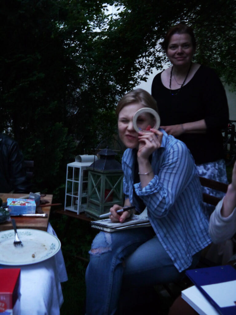Zwei Menschen schauen in die Kamera, sitzenund stehen im Garten. Eine schaut durch eine Klebeben-Rolle und hält Skizzenbuch, sowie Stift in der Hand