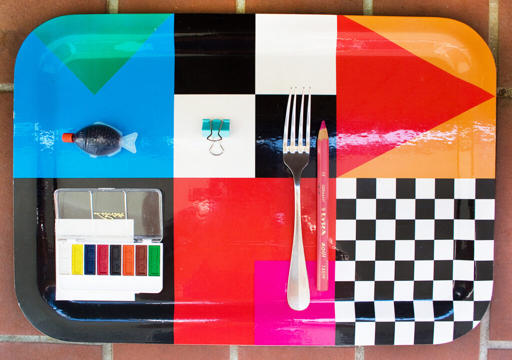 Blick von oben auf ein buntes Tablett: Aquarellfarbe, Sojasauce, Klammer, Gabel, Buntstift geometrisch angeordnet