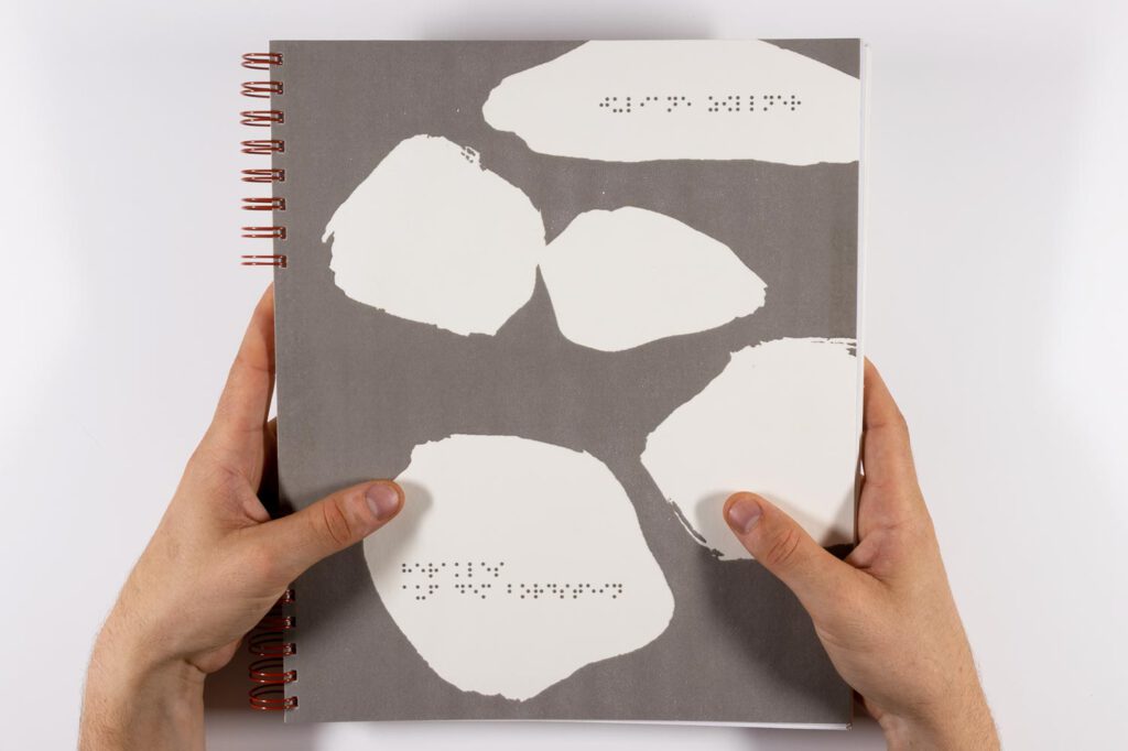 Zwei Hände halten ein Buch mit Braille-Schrift und Schwellpapier-Illustration in den Händen. Der Text lautet: Juliane Zöllner, Herakles auf dem Bordstein.