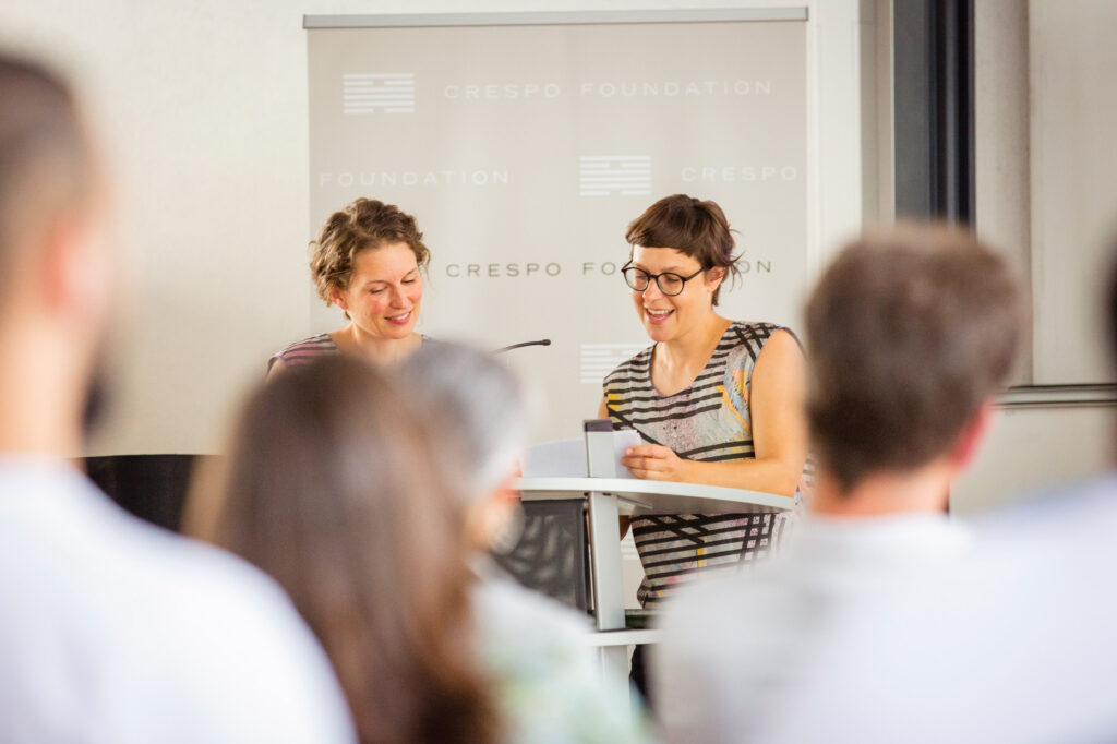 Marina Kampka (links im Bild) und Lena Schrieb (rechts im Bild) sprechen vor Publikum in ein Mikrofon an einem Redner:innenpult. 