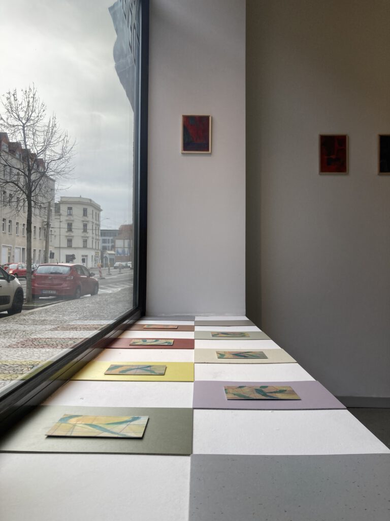 Zeichnungen und Linoleum liegen im Schaufenster des Kunstraum Modos Dever in Leipzig. Links der Blick durch das Schaufenster auf die Straße mit stehenden Autos, rechts der Blick auf einige Bilder im Ausstellungsraum.